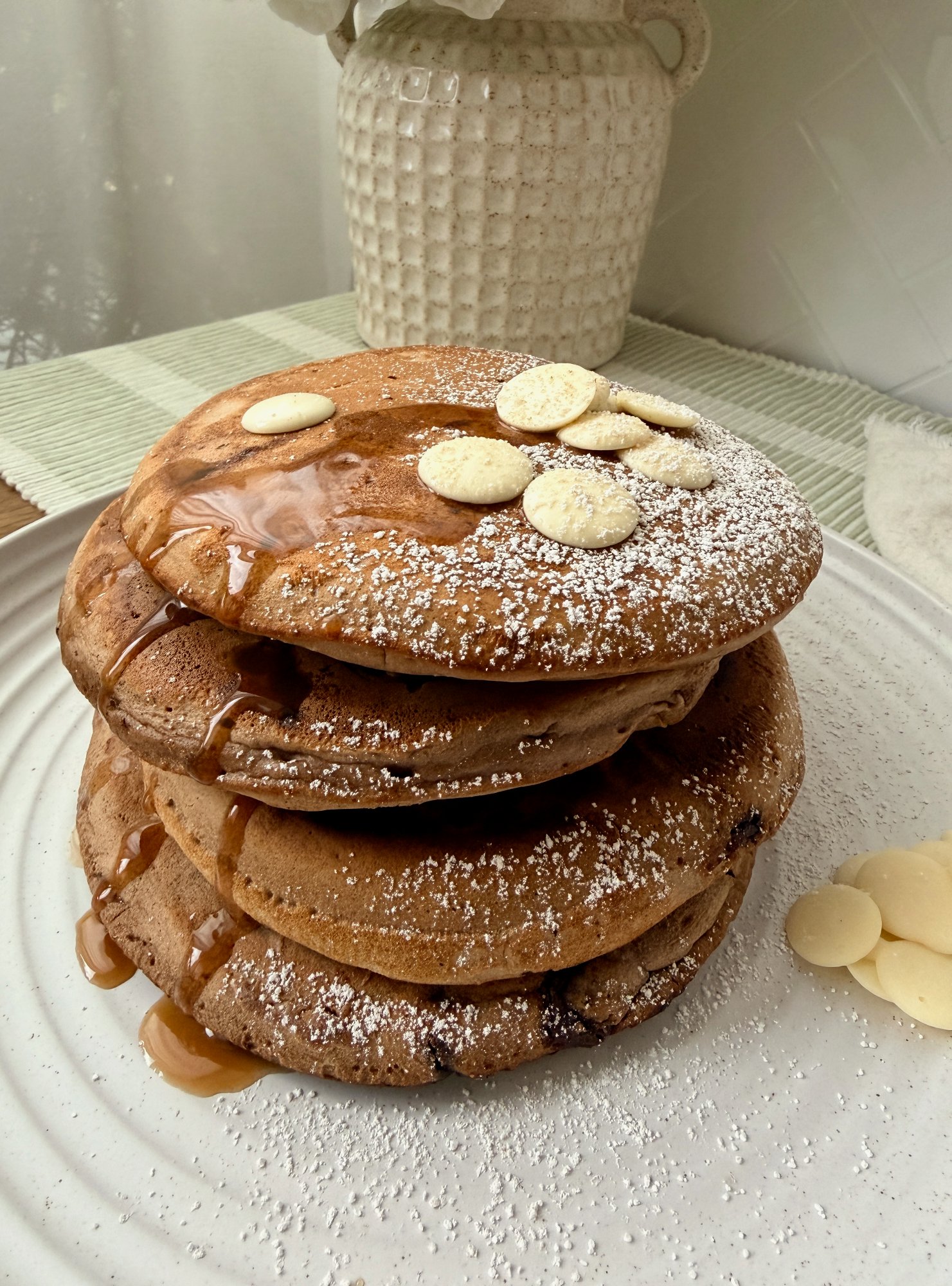 A stack of thick double chocolate pancakes with maple syrup, icing sugar and white chocolate buttons.