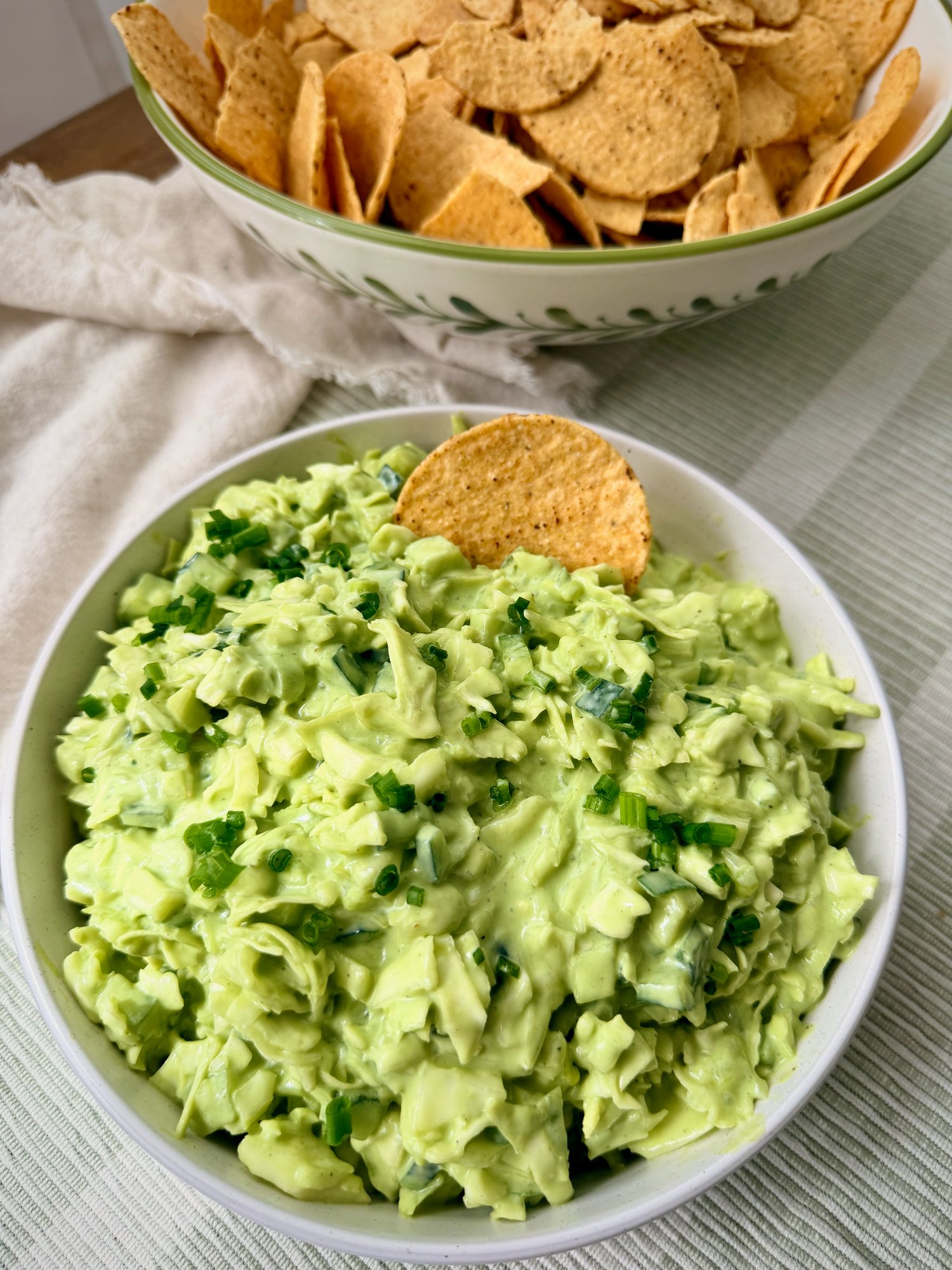 A white bowl of finely chopped green goddess cabbage salad scattered with spring onion, a corn chip standing upright in the centre, with a bowl of corn chips behind.