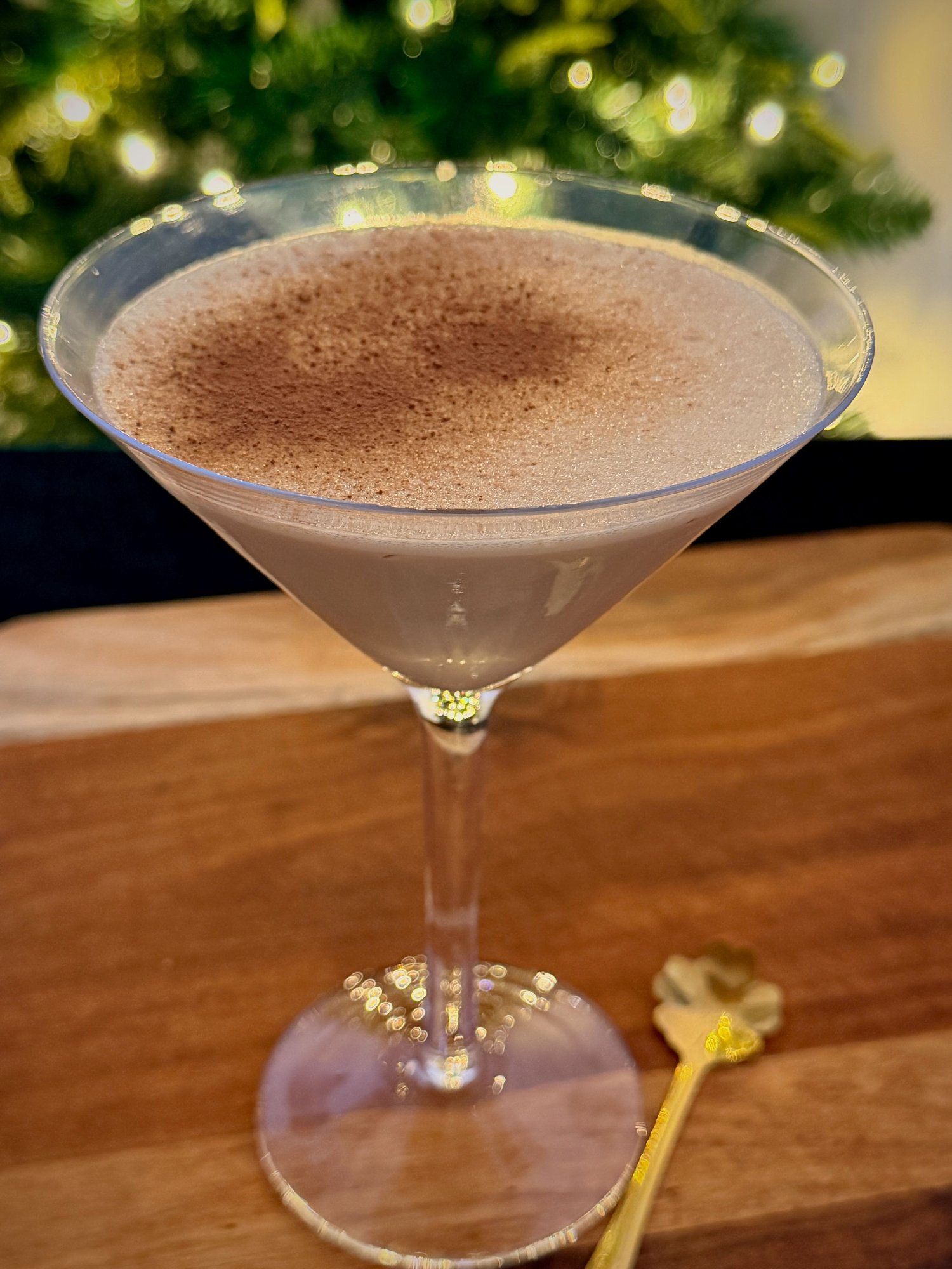 Tiramisu martini with a light cocoa dusting in a martini glass, with a gold spoon and Christmas lights behind.