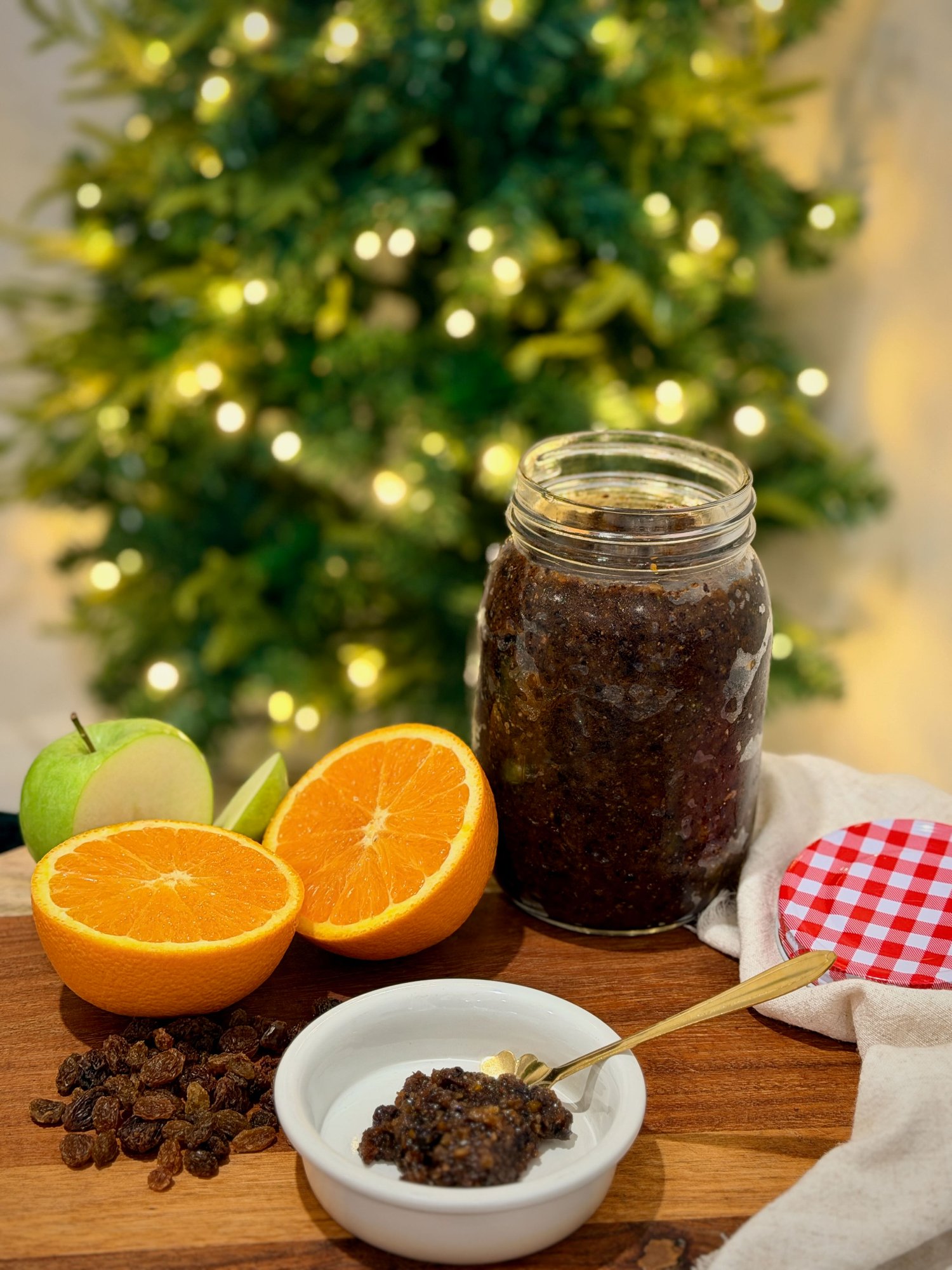Homemade fruit mince in a glass jar with orange halves, sliced green apple and sultanas on a timber board.