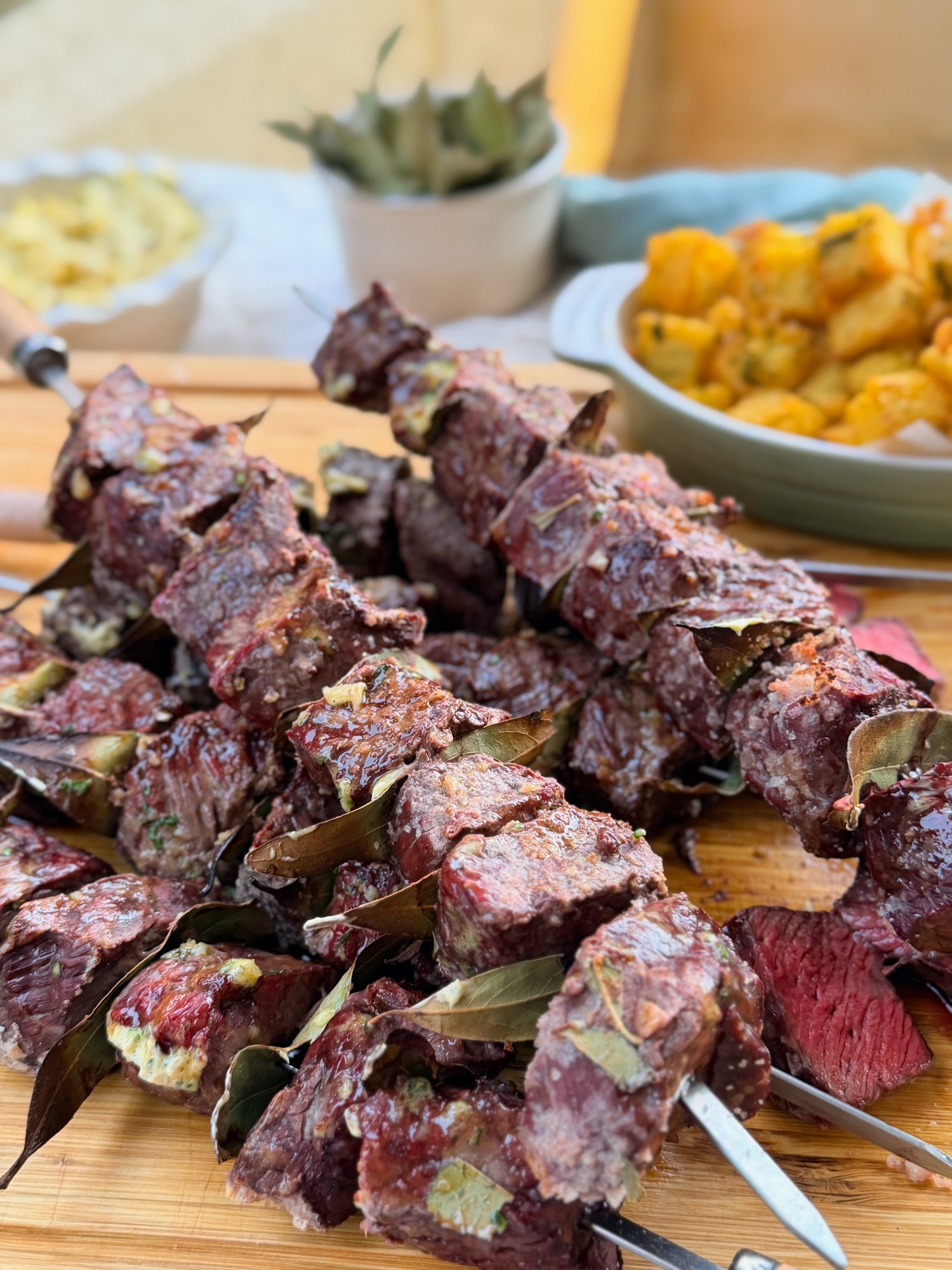 Portuguese espetada skewers brushed with garlic butter on a board, with Milho Frito, garlic butter and bay leaves behind.