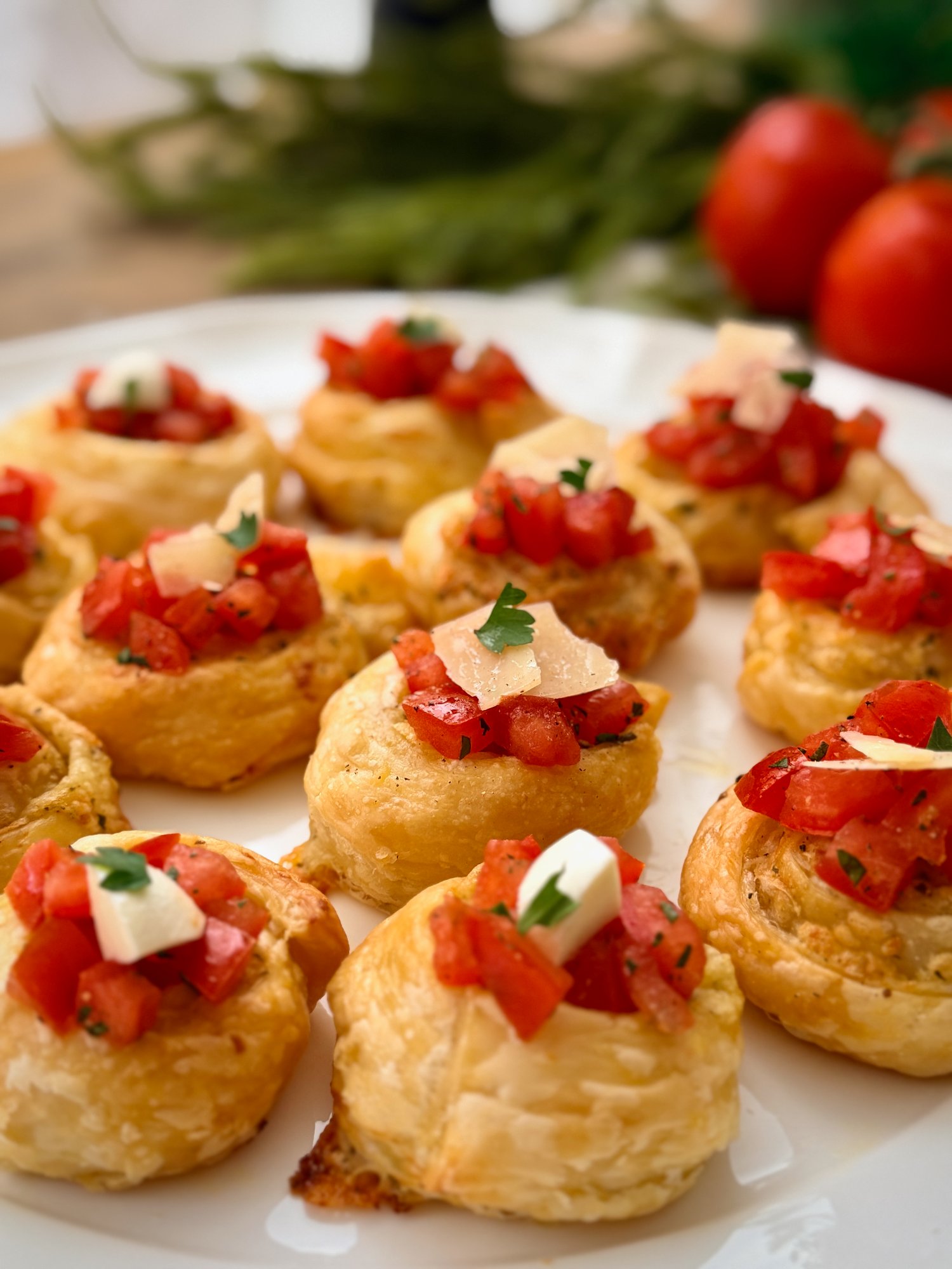Garlic parmesan bruschetta bites on a white platter, golden puff pastry scrolls topped with diced tomato, shaved parmesan, bocconcini and parsley, with vine tomatoes behind.
