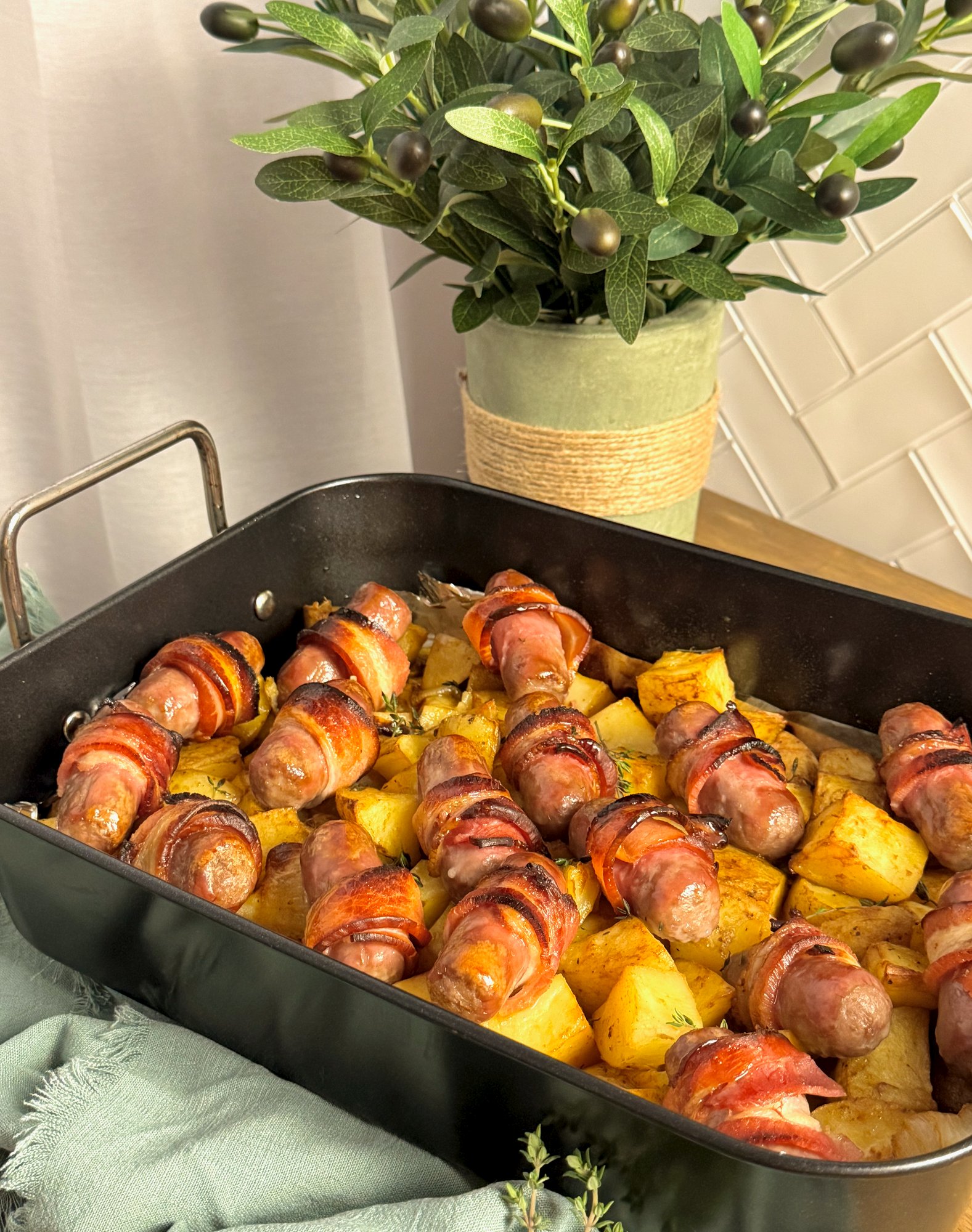 A tray of crispy bacon wrapped sausages roasted over golden potatoes, onion and garlic.